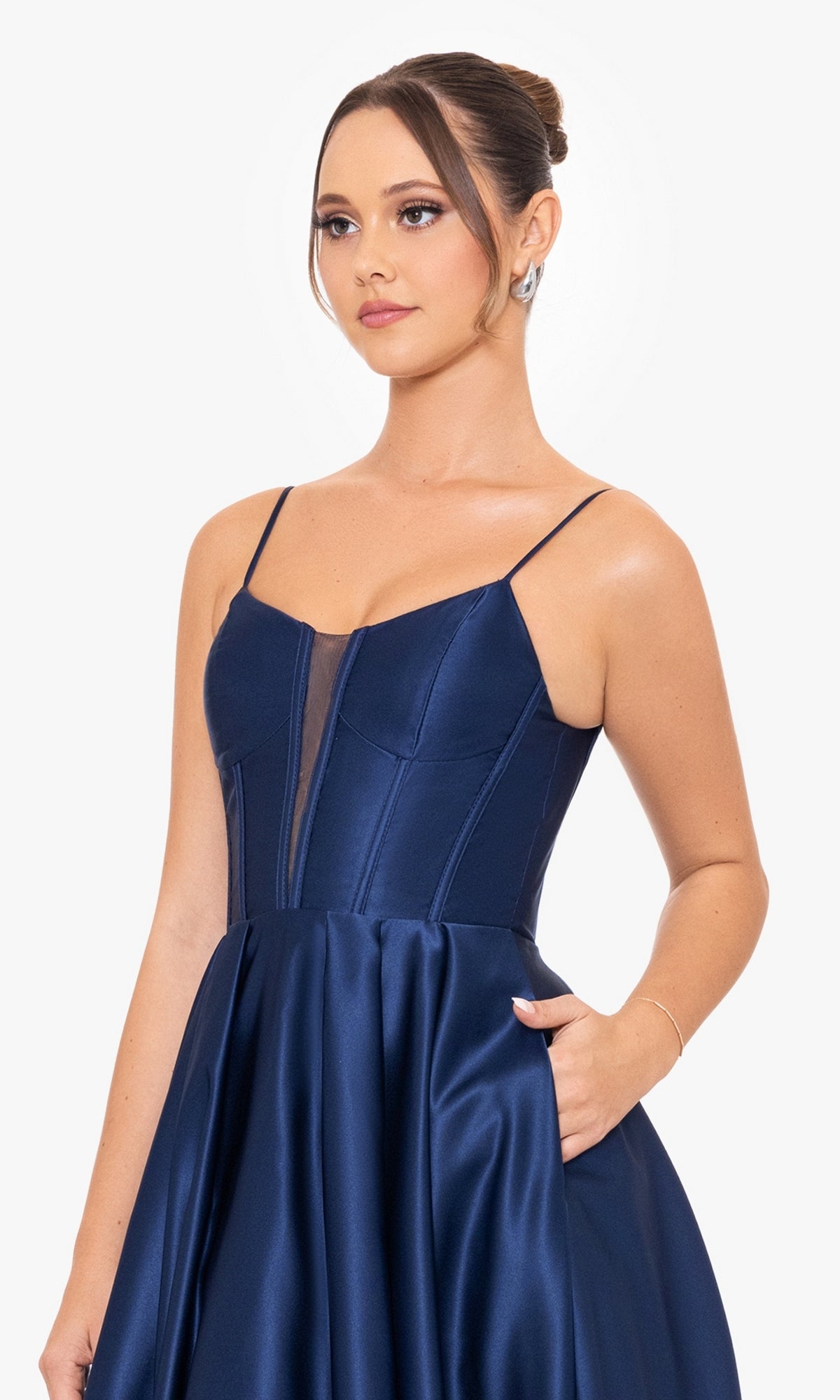 A woman in the Blondie Nites Long A-Line Prom Dress 4826BN, a sleeveless navy blue satin gown with thin straps and a sheer corset-style bodice, poses with her hand in her pocket against a white background.